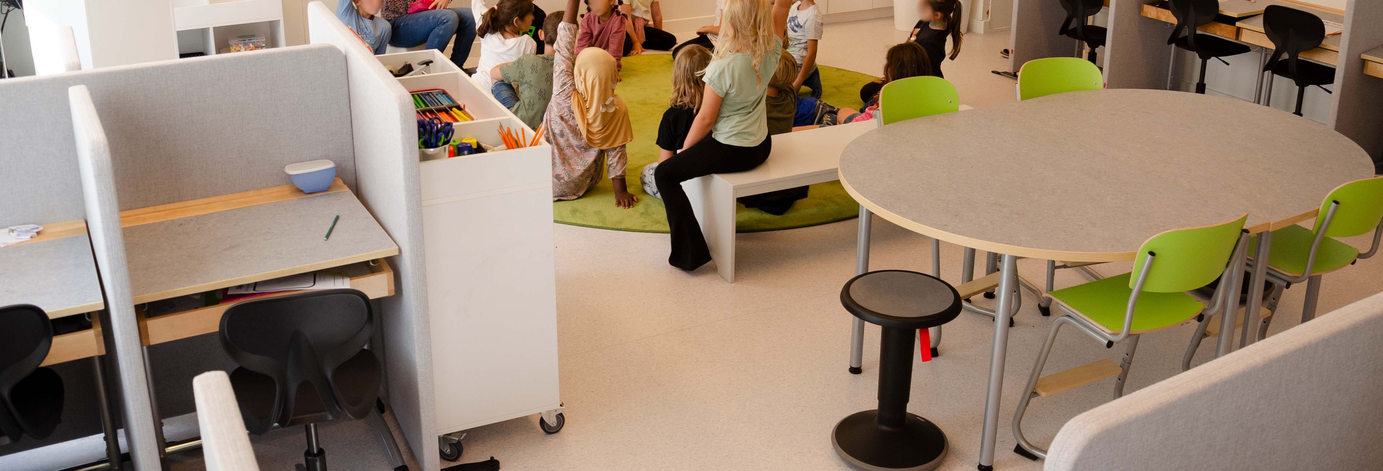 Här har barnen samling på den stora, gröna, runda mattan. Här syns också det ovala bordet i mitten av klassrummet med gröna stolar och en balanspall.